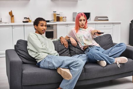 Foto de Sorprendido hermosa pareja multirracial sentado en el sofá y ver películas en la sala de estar en casa - Imagen libre de derechos