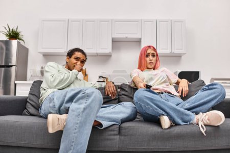 verängstigtes multikulturelles Paar in kuscheliger Hauskleidung, das zu Hause im Wohnzimmer Horrorfilme guckt