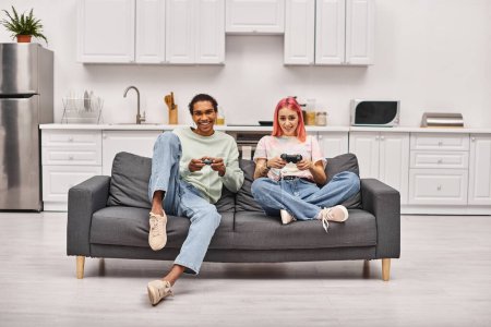 cheerful interracial couple in homewear sitting on sofa and playing games using gamepads at home
