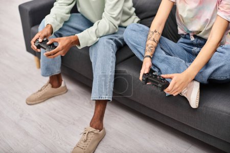 Foto de Vista recortada de joven pareja multicultural en atuendos casuales jugando juegos con gamepads en casa - Imagen libre de derechos