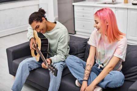 Foto de Alegre rosa peludo mujer viendo su guapo alegre novio jugando guitarra mientras en casa - Imagen libre de derechos