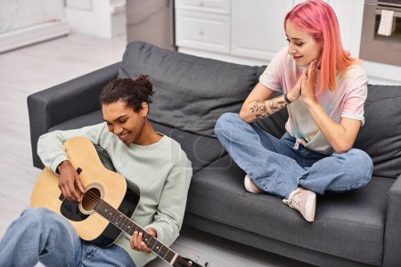 Foto de Mujer alegre con pelo rosa mirando a su alegre novio afroamericano tocando la guitarra - Imagen libre de derechos