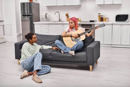 Foto de Atractiva pareja diversa en ropa de casa mirándose mientras están sentados en el sofá, la guitarra en la mano - Imagen libre de derechos