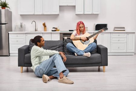 Foto de Alegre afroamericano hombre en ropa de casa mirando amorosamente a su novia de pelo rosa tocando la guitarra - Imagen libre de derechos