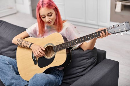Foto de Atractiva mujer alegre con el pelo rosa sentado en el sofá y tocando la guitarra mientras está en la sala de estar - Imagen libre de derechos