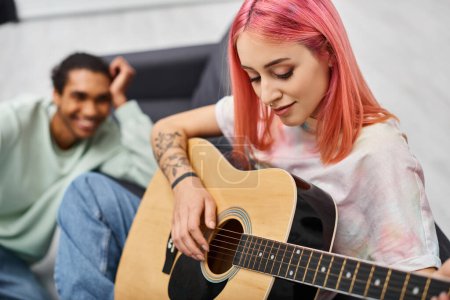 Foto de Enfoque en la mujer de pelo rosa tocando la guitarra delante de su novio afroamericano alegre borrosa - Imagen libre de derechos