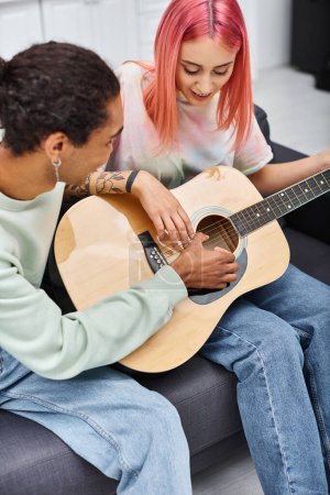Foto de Buen aspecto alegre afroamericano hombre enseñando a su novia de pelo rosa cómo tocar la guitarra - Imagen libre de derechos