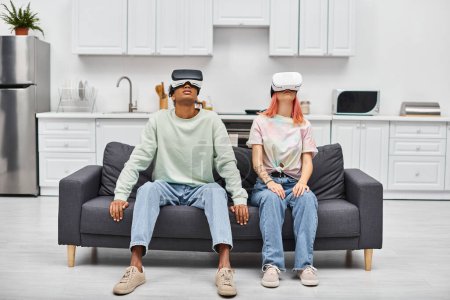 gut aussehendes multiethnisches Paar in gemütlicher Homewear, das zu Hause auf dem Sofa sitzt und VR-Headsets trägt