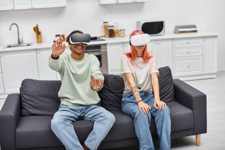 fröhlich ansprechendes multikulturelles Paar sitzt mit VR-Headsets auf Sofa im Wohnzimmer