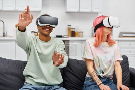 alegre hermosa pareja multirracial en ropa de casa sentado en el sofá con auriculares de realidad virtual