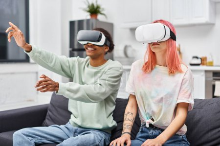 Photo for Joyful attractive diverse couple in casual attire sitting on sofa in living room with VR headsets - Royalty Free Image