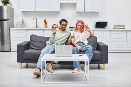 Téléchargez les photos : Joyeux beau couple multiculturel assis sur le canapé et agitant à la caméra de l'ordinateur portable pendant l'appel vidéo - en image libre de droit