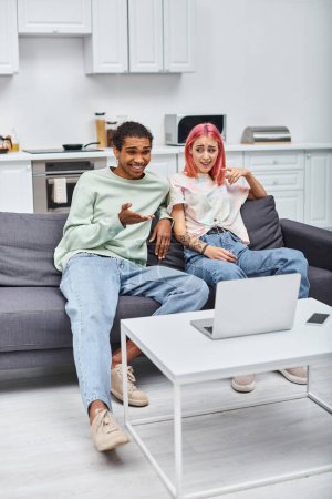 Foto de Alegre interracial pareja en homewear teniendo video llamada y sentado en sofá en sala de estar - Imagen libre de derechos