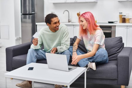 joyous multiracial couple in cozy attire holding credit card near laptop and smiling at each other