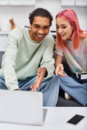 Foto de Atractiva pareja multicultural alegre mirando portátil con tarjeta de crédito en la mano mientras está en casa - Imagen libre de derechos
