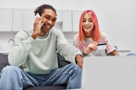 Foto de Atractiva pareja alegre diversa mirando el ordenador portátil mientras sostiene la tarjeta de crédito y hablando por teléfono - Imagen libre de derechos