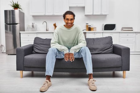 Téléchargez les photos : Joyeux jeune homme afro-américain dans des vêtements confortables assis sur le canapé et regardant la caméra à la maison - en image libre de droit