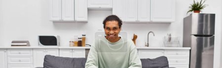 homme afro-américain joyeux en tenue décontractée sur canapé dans le salon souriant à la caméra, bannière