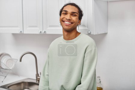 Foto de Alegre atractivo afroamericano hombre en suéter blanco cálido sonriendo felizmente a la cámara en casa - Imagen libre de derechos