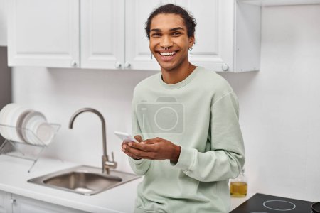 attrayant homme afro-américain joyeux dans des vêtements confortables tenant smartphone et souriant à la caméra