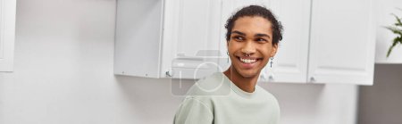 good looking cheerful african american man in green sweater looking away while in kitchen, banner