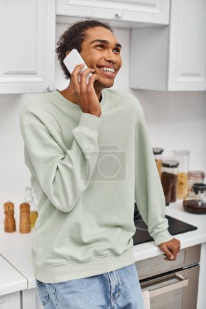 Téléchargez les photos : Beau beau homme afro-américain joyeux en pull vert parlant joyeusement par téléphone et détournant les yeux - en image libre de droit