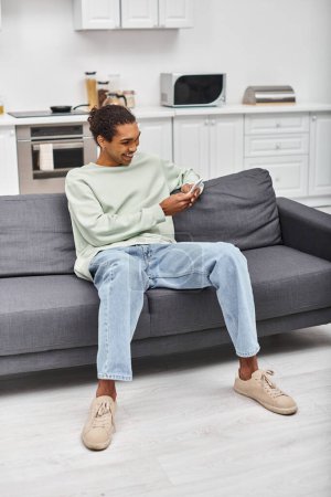 guapo alegre afroamericano hombre en cómoda ropa de casa sentado en el sofá y mirando el teléfono