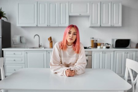 Foto de Atractiva mujer joven con el pelo rosa en acogedora ropa de casa sentado en la mesa y mirando a la cámara - Imagen libre de derechos