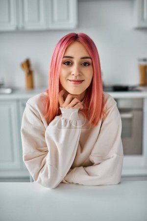 Foto de Alegre joven con el pelo rosa vibrante sentado y sonriendo a la cámara mientras que en la cocina en casa - Imagen libre de derechos