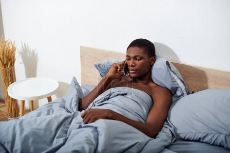 An African American man lays in bed, chatting on his cell phone in the morning, starting his day off with a phone call.