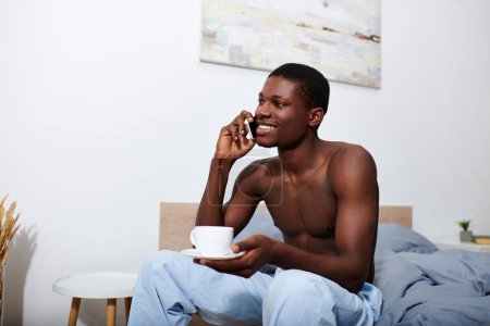 Young African American man sits on bed in morning, engrossed in conversation on cell phone.