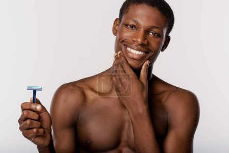 Homme afro-américain torse nu dans un studio avec un fond blanc, engagé dans sa routine beauté, tenant un rasoir dans sa main droite.