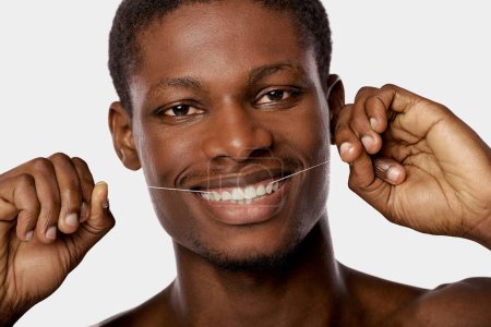 Smiling shirtless African American man playfully flosses his teeth with a string in a studio setting.