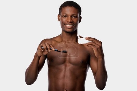 A shirtless African American man holds a toothbrush and toothpaste in his right hand against a white background.