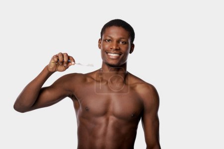 Hombre afroamericano sin camisa en un estudio sosteniendo un cepillo de dientes en su mano derecha sobre un fondo blanco.