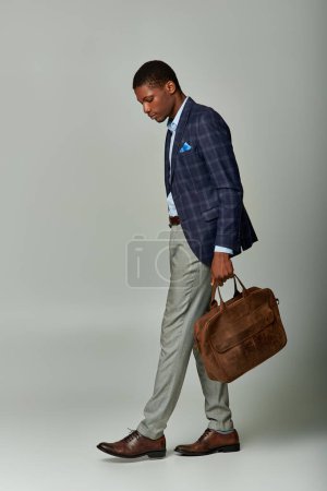 Schöner afroamerikanischer Geschäftsmann im karierten Blazer geht selbstbewusst mit Aktentasche in der Hand auf grauem Hintergrund.