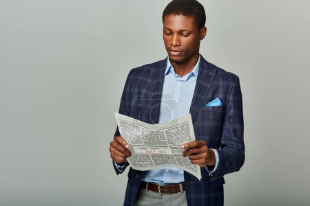 Schöner afroamerikanischer Geschäftsmann im karierten Blazer, in Zeitung vertieft auf grauem Hintergrund.