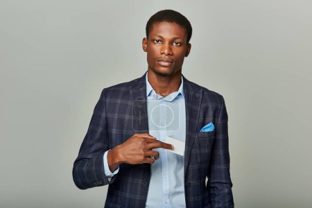 Handsome African American businessman in checkered blazer confidently holds up a business card against a grey background.