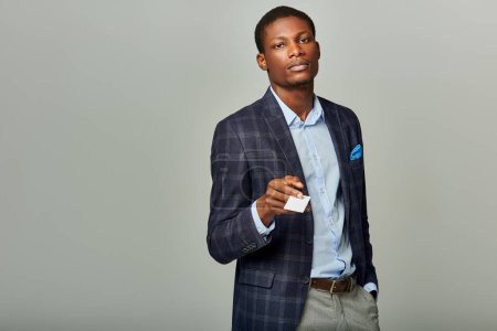 A dashing African American businessman in a checkered blazer holds out a business card against a grey background.