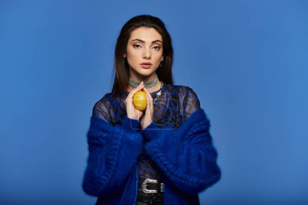 A young woman in a blue sweater holds a lemon in front of a blue background.