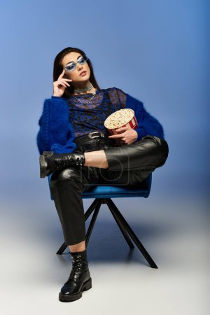 A stylish woman sits in a blue chair, holding a bucket of popcorn.