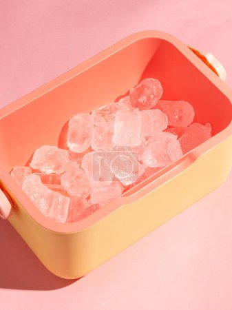Recipiente rosa y amarillo lleno de cubitos de hielo sobre un fondo rosa suave. Presenta un diseño minimalista y moderno, perfecto para temas relacionados con el verano, la frescura y las bebidas frescas.