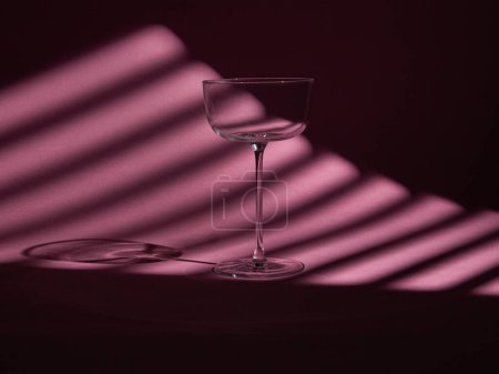 A clear wine glass stands against a backdrop of dramatic, striped light and shadow. The interplay of light creates a moody and artistic ambiance, perfect for themes of elegance, minimalism, and modern design.
