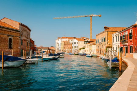 Murano, Italy - October 2022: Murano Canal with colorful houses, moored boats and cranes in the background