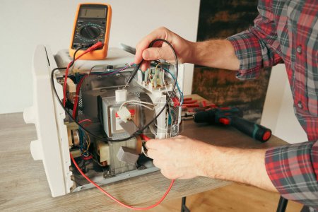 Photo for A repair engineer uses a multimeter to check the circuits of a microwave oven, looking for a fault. Diagnostics repair of microwave oven. - Royalty Free Image