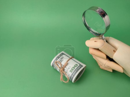 Wooden hand holding magnifying glass with banknotes on a green background.