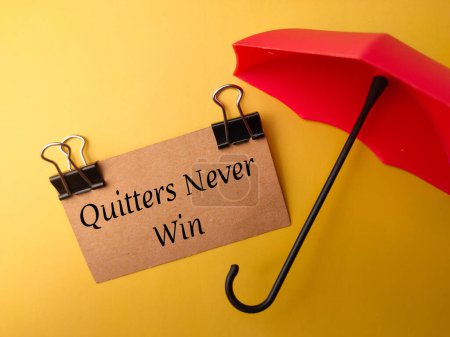 Regenschirm und braune Karte mit Text Quitters Never Win auf gelbem Hintergrund.
