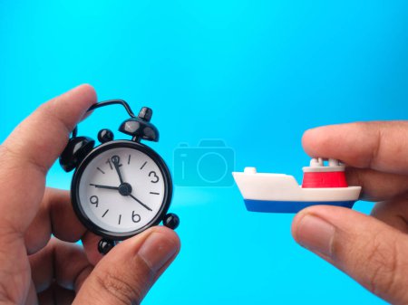 Téléchargez les photos : Main tenant horloge et jouets navires sur fond bleu. Concept de voyage. - en image libre de droit