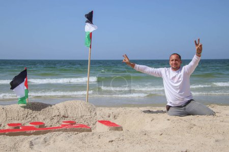 Téléchargez les photos : L'artiste palestinien Muhammad Totah avec sa jambe amputée peint sur la sculpture de sable Open Gazas points de vente. 28 septembre 2023. Gaza, Palestine - en image libre de droit