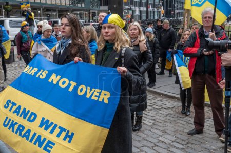 Photo for Mayor Adams Delivers Remarks At Flag-Raising Ceremony For Ukraine. February 24, 2024, New York, New York, USA: Women hold a WAR IS NOT OVER STAND WITH UKRAINE sign at a Flag-Raising Ceremony for Ukraine - Royalty Free Image